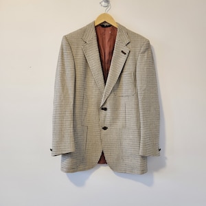Puede incluir: Un blazer a cuadros beige y negro con forro interior marrón. El blazer tiene dos botones negros y un pequeño bolsillo en el lado izquierdo. Está colgado de una percha de madera.