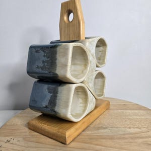 May include: A ceramic mug holder with four stacked, open-ended mugs in shades of blue and cream. The mugs sit on a wooden base with a wooden handle. The mugs have a unique, geometric shape.