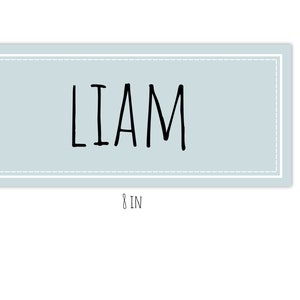 Op de afbeelding: Een lichtblauw rechthoekig bord met een witte rand en witte stiksels rond de rand. Het bord zegt "LIAM" in zwarte handgeschreven letters. Het bord is 20,3 cm breed en 8,9 cm hoog.
