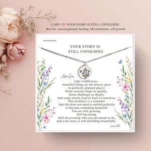 May include: A silver necklace with a round pendant featuring a floral design, presented on a card with the text "YOUR STORY IS STILL UNFOLDING". The card includes a message about growth and self-discovery, with floral illustrations.