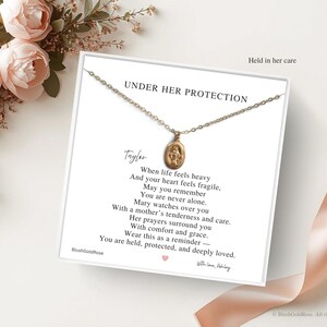 May include: A gold-colored necklace with a pendant featuring a religious image, displayed on a card with the text "UNDER HER PROTECTION" and a poem. The image is set against a white background with floral accents and a ribbon.