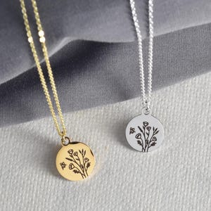 May include: Two delicate necklaces with round pendants. One is gold-toned, the other silver-toned. Each pendant features a floral design with a small butterfly. The necklaces are displayed on a textured, light gray surface.