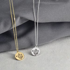 May include: Two delicate necklaces with round pendants. One is gold-toned, the other silver-toned. Each pendant features a floral design. The necklaces hang against a gray fabric background, showcasing the jewelry's details and design.