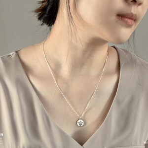 May include: A silver box chain necklace with a round pendant featuring a floral design. The necklace is shown on a person wearing a beige top. The words "WILD FLOWERS" are visible in the image.