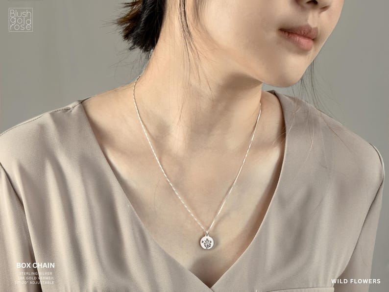 May include: A silver box chain necklace with a round pendant featuring a floral design. The necklace is shown on a person wearing a beige top. The words "WILD FLOWERS" are visible in the image.