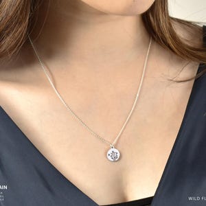 May include: A silver cable chain necklace with a small, round pendant featuring a wildflower design. The necklace is shown on a person wearing a dark blue top. The words "Wild Flowers" are visible.