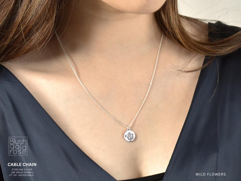 May include: A silver cable chain necklace with a small, round pendant featuring a wildflower design. The necklace is shown on a person wearing a dark blue top. The words "Wild Flowers" are visible.