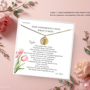 May include: A gold-colored necklace with a circular pendant featuring a floral design, presented on a card with inspirational text. The card is surrounded by pink flowers and greenery, creating a soft, feminine aesthetic.