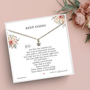 May include: A delicate silver necklace with a star pendant, presented on a white card with floral accents. The card reads "KEEP GOING" and includes an inspirational message. The background features pink flowers.