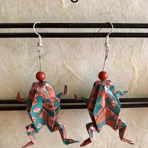 May include: A pair of origami frog earrings with a floral pattern in shades of pink, green, and brown. The earrings are made of paper and have silver hooks.