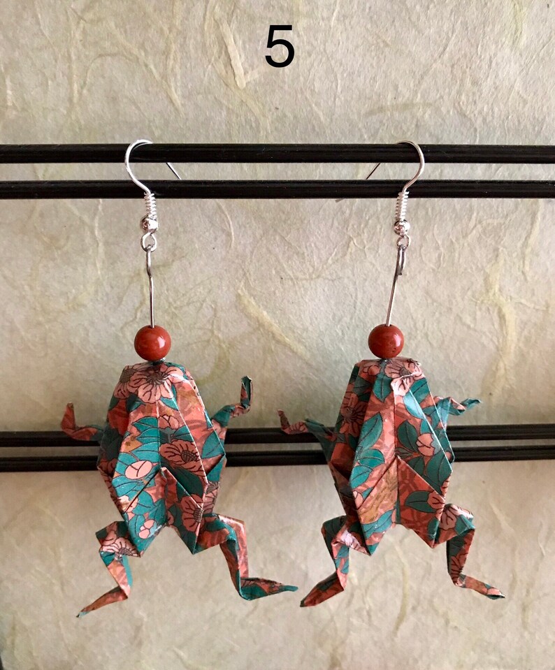 May include: A pair of origami frog earrings with a floral pattern in shades of pink, green, and brown. The earrings are made of paper and have silver hooks.