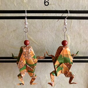 May include: A pair of origami frog earrings with a green, brown, and gold patterned design. The earrings are hanging from silver hooks with brown beads.