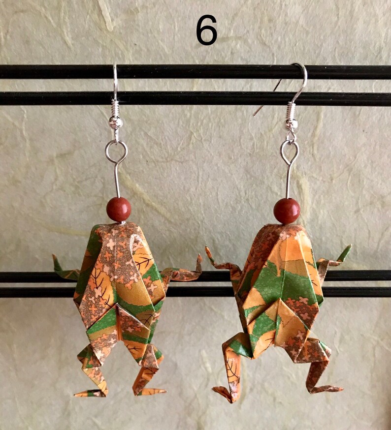 May include: A pair of origami frog earrings with a green, brown, and gold patterned design. The earrings are hanging from silver hooks with brown beads.