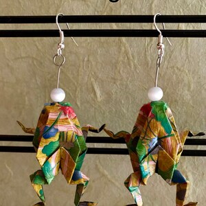 May include: A pair of origami frog earrings with a floral pattern and white pearl accents. The earrings are made of paper and have silver hooks.