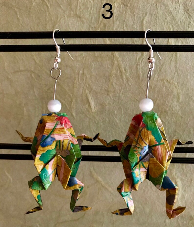 May include: A pair of origami frog earrings with a floral pattern and white pearl accents. The earrings are made of paper and have silver hooks.