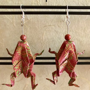 May include: A pair of origami frog earrings with a red and green patterned paper design. The earrings are hanging from silver hooks with a brown bead at the top of each hook.