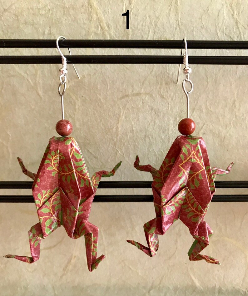 May include: A pair of origami frog earrings with a red and green patterned paper design. The earrings are hanging from silver hooks with a brown bead at the top of each hook.