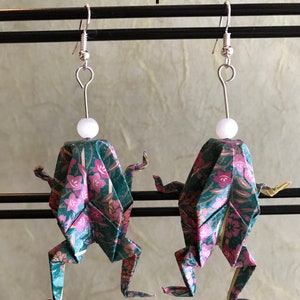 May include: A pair of origami frog earrings with a green and pink floral pattern. The earrings are made of paper and have a white bead at the top of each earring.