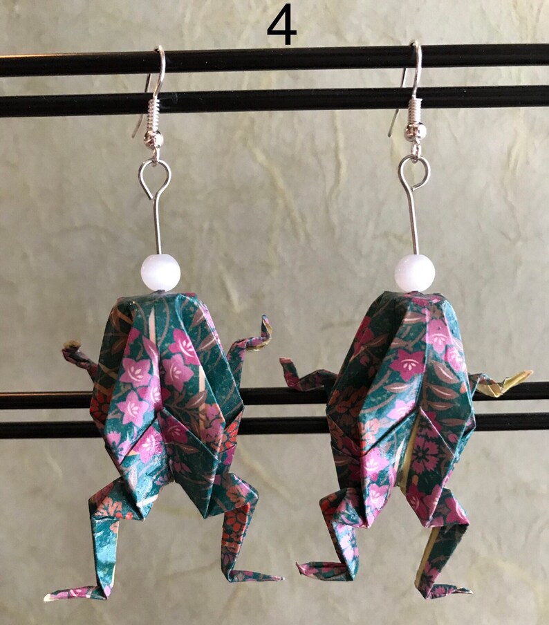 May include: A pair of origami frog earrings with a green and pink floral pattern. The earrings are made of paper and have a white bead at the top of each earring.