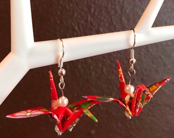May include: A pair of red origami crane earrings with white pearl accents. The earrings are hanging from silver hooks.