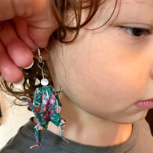 May include: A pair of origami frog earrings with a floral pattern on a green background. The earrings are made of paper and have a white bead at the top.