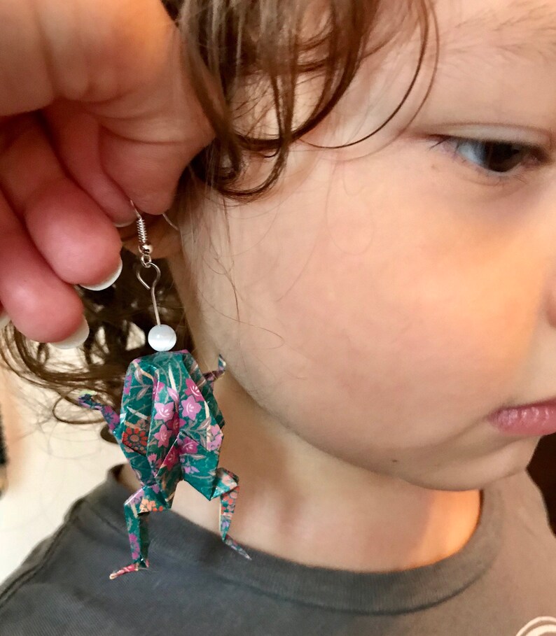 May include: A pair of origami frog earrings with a floral pattern on a green background. The earrings are made of paper and have a white bead at the top.