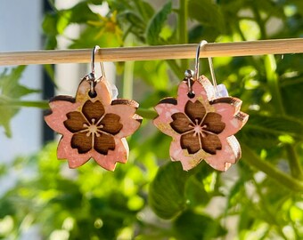 May include: A pair of pink and gold cherry blossom earrings made of wood. The earrings are hanging from a wooden stick.