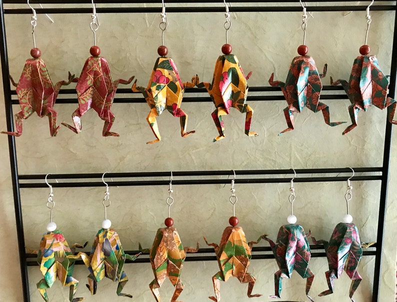 May include: A display of 10 pairs of origami frog earrings. The frogs are made of patterned paper and have a bead at the top of their heads. The earrings are hanging from a black metal rack.