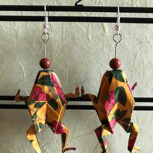 May include: A pair of origami frog earrings with a yellow, green, and red floral pattern. The earrings are hanging from silver hooks with brown beads.