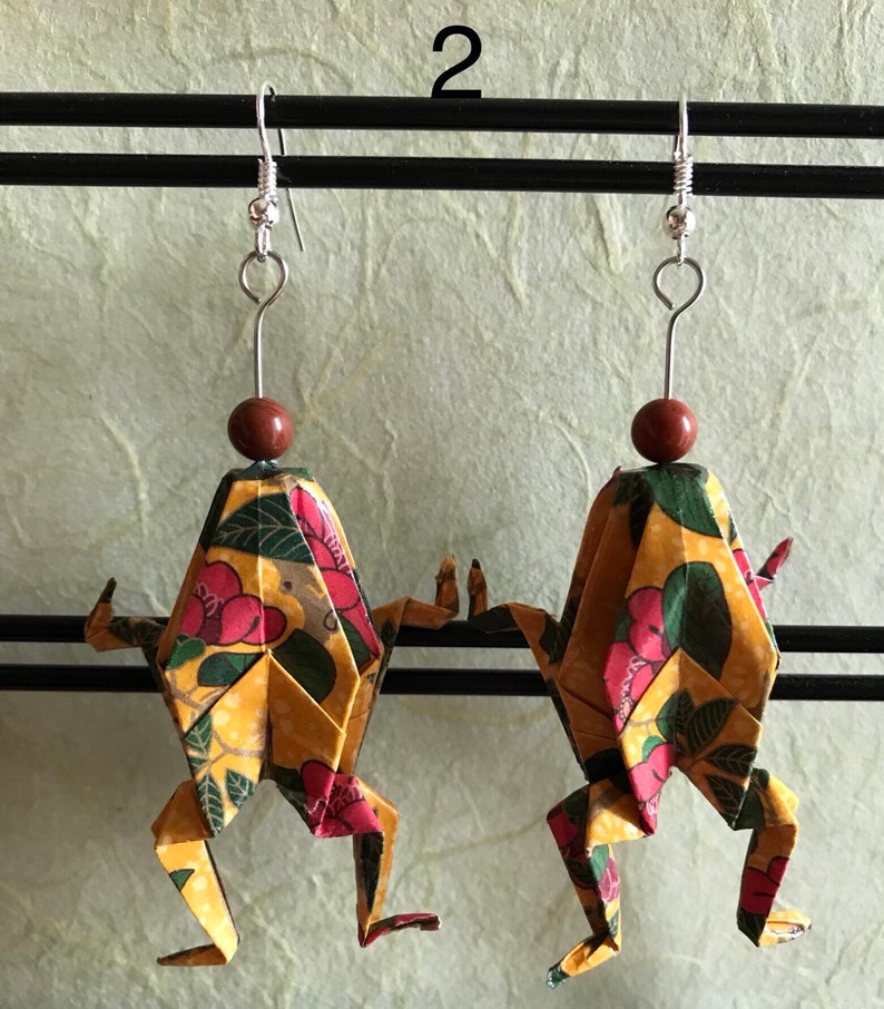 May include: A pair of origami frog earrings with a yellow, green, and red floral pattern. The earrings are hanging from silver hooks with brown beads.