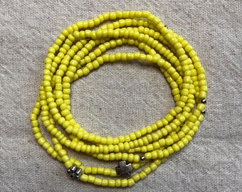 May include: A coiled strand of yellow beads with silver-colored accents and a cross-shaped charm. The beads are small and evenly spaced, creating a delicate, layered appearance. The background is a textured, neutral-toned fabric.