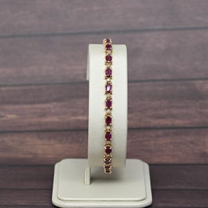 May include: A gold tennis bracelet featuring a series of oval-cut deep red rubies alternating with small clear round-cut diamonds. The gemstones are set in a continuous metal chain design, showcasing a vibrant contrast between the red and gold.