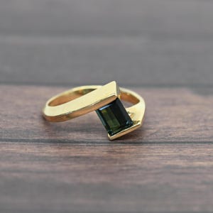 May include: A gold ring with a rectangular, dark green gemstone. The ring has a modern design with a thick band and a geometric setting for the gemstone. The ring is displayed on a wooden surface.