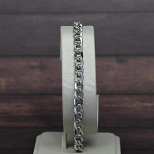 May include: A silver-toned curb chain bracelet featuring interlocking oval links. Each individual link displays a polished finish on the outer edges and a textured, cross-hatched pattern on the inner surface, creating a reflective metallic appearance.