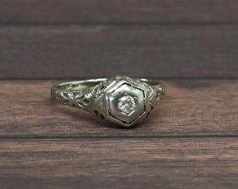 May include: A silver ring with a hexagonal setting and a small, clear gemstone at the center. The ring has intricate filigree detailing on the band and around the setting. The ring is displayed on a dark wood surface.