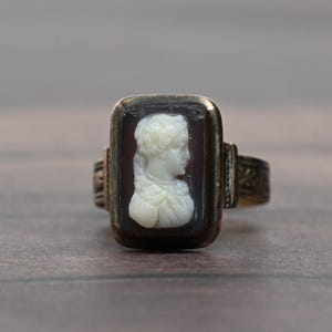 May include: A rectangular ring featuring a carved white cameo of a profile portrait set against a dark brown stone. The ring has a metallic band with decorative etched patterns on the sides, framing the central cameo in a polished metal setting.
