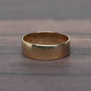 May include: A gold band ring with a smooth, polished exterior surface. The interior of the ring features a visible engraved inscription in cursive script, showing a name or word that is partially obscured and difficult to read clearly.