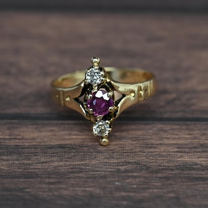 May include: A gold ring with a central, round, deep pink gemstone, flanked by two smaller, clear gemstones. The ring has an ornate design with a textured band. The ring is set against a dark wood grain background.