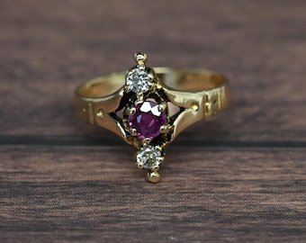 May include: A gold ring with a central, round, deep pink gemstone, flanked by two smaller, clear gemstones. The ring has an ornate design with a textured band. The ring is set against a dark wood grain background.