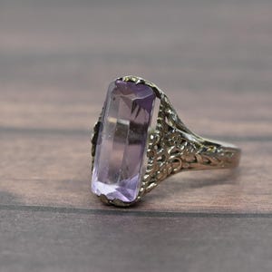 May include: A silver-toned ring featuring a large, rectangular, faceted light purple gemstone set in a decorative, openwork metal band with intricate floral-inspired filigree detailing along the sides of the setting and the shank.