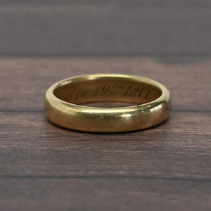 May include: A smooth, polished gold band ring with a rounded profile. The interior surface of the ring features a clear, dark engraved inscription that reads Nov 19th 1867, showcasing a classic design with a visible metallic luster and finish.