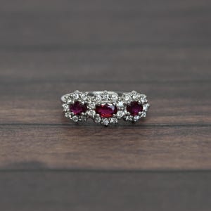 May include: A silver ring featuring three oval-cut, deep red gemstones, each surrounded by a halo of smaller, clear stones. The ring is set against a dark wood grain background, highlighting the sparkle of the gems.