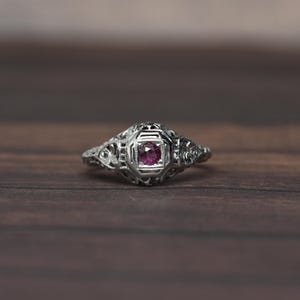 May include: A silver ring with a square, deep pink gemstone at the center. The ring has an ornate, filigree design around the gemstone and along the band. The ring is set against a dark wood surface.