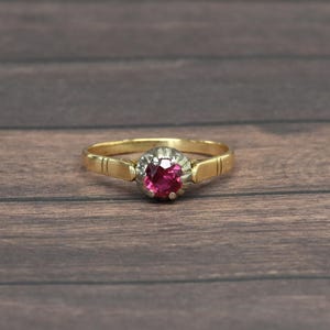 May include: A gold ring with a round, bright red gemstone at the center. The gemstone is surrounded by a silver-colored setting. The ring band has a simple design with etched lines. The ring is set against a dark wood background.