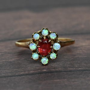 May include: A gold ring featuring a central oval-cut deep red gemstone surrounded by a halo of eight small, round, iridescent white opals. The stones are set in a pronged metal setting on a plain, polished gold band with a smooth, rounded finish.