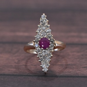 May include: A gold ring featuring a central, round, deep red gemstone surrounded by numerous smaller, clear gemstones. The ring's design is elongated, with the gemstones arranged in a tapering, vertical pattern. The ring is set against a dark wood grain background.