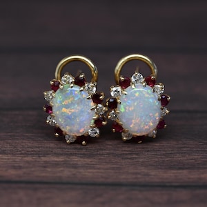 May include: A pair of gold earrings featuring large, iridescent opal cabochons surrounded by a halo of small, red rubies and clear, sparkling diamonds. The earrings have a post and hoop design, set against a dark wood grain background.
