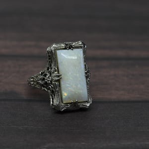 May include: A rectangular white opal gemstone ring with an ornate, filigree silver setting. The opal has a subtle play of color. The ring is set against a dark wood grain background, highlighting the jewelry's details.