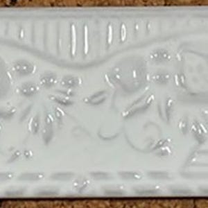 May include: A white ceramic tile featuring a decorative floral and fruit design. The tile is rectangular with pointed ends. The embossed design includes flowers, leaves, and fruit, adding a touch of elegance.