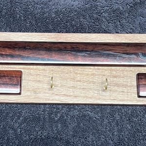 May include: Wooden key holder with the word "Jeep" engraved in the center. The key holder has a light brown base with two dark brown rectangular accents and a dark brown shelf. It features four brass hooks for keys.